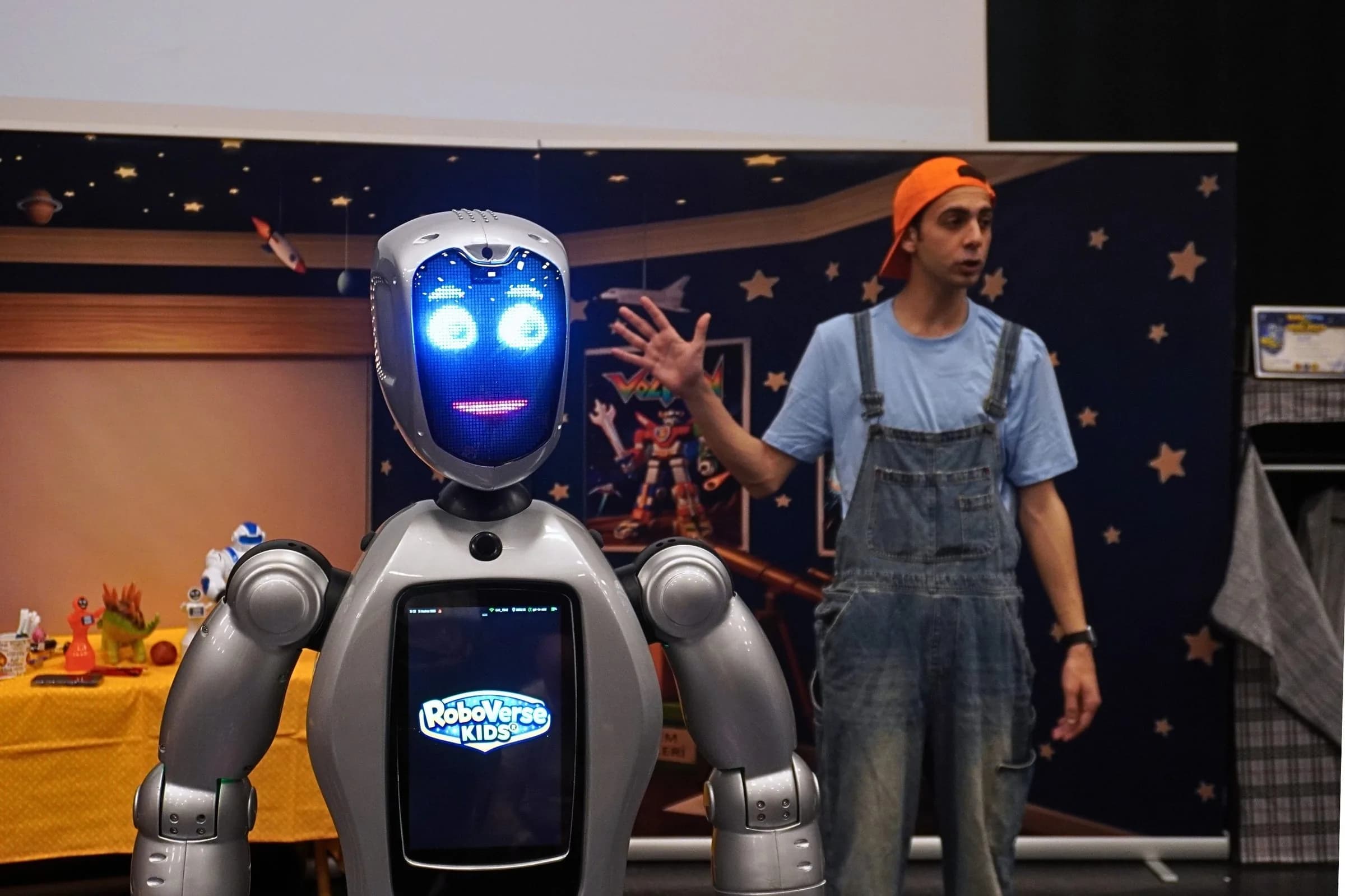 Sahnede gerçek robot ve tulumlu bir oyuncu; arkada yıldızlı lacivert duvarlı dekor, robotun göğüs ekranında RoboVerse Kids logosu